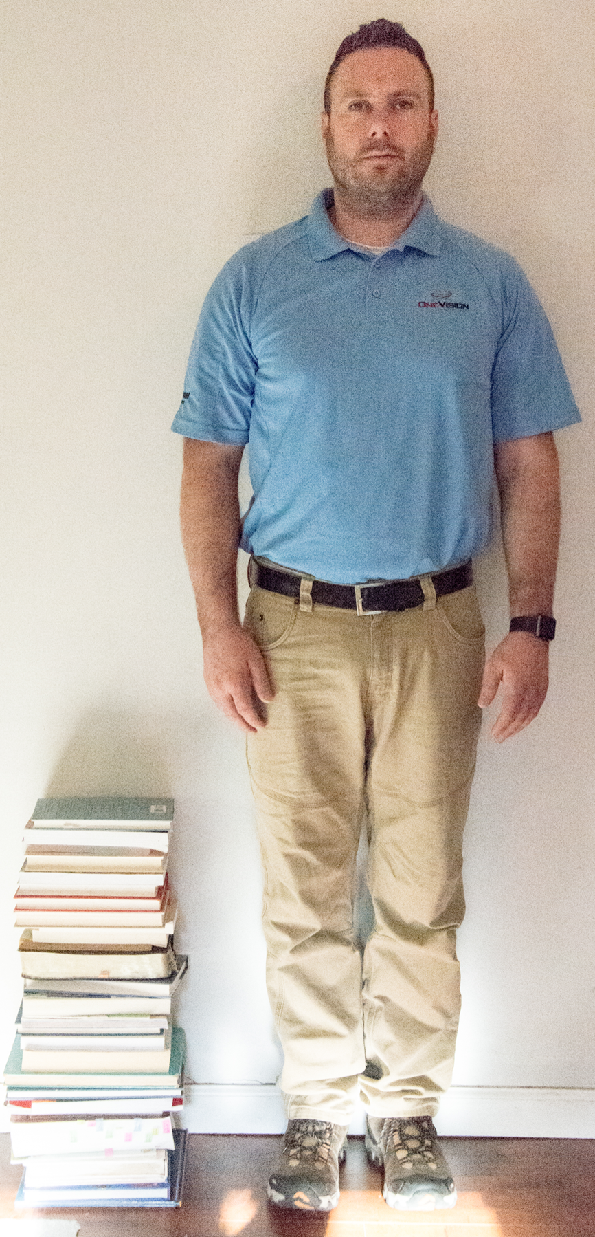 Image of stack of books next to adult man. Stack reaches man's middle thigh.