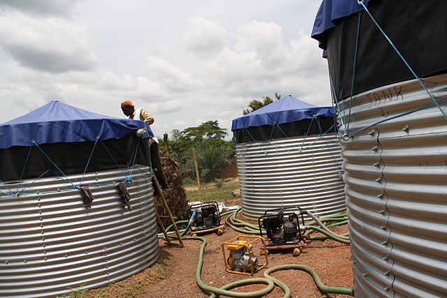 Photo of three water tanks with drainage hoses. A worker is on a ladder, leaned against the side of one of the tanks.