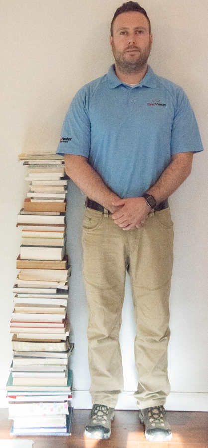 Image of stack of books next to adult man. Stack reaches man's elbow.