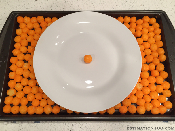 A picture of a tray with covered with one layer of circular cheese puffs. On top of the cheese puffs is a circular plate with 1 cheese puff placed in the center of the plate.